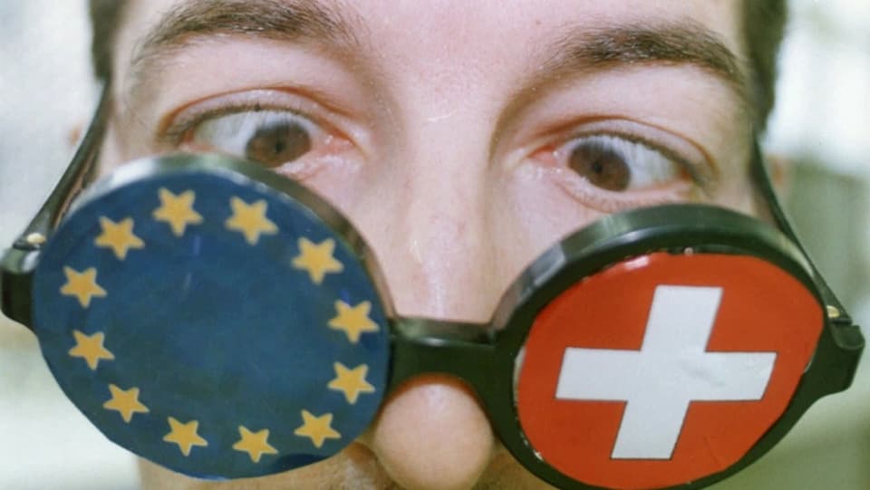 Ein Mann schielt durch eine Brille, auf deren einem Glas ein Schweizerkreuz, auf dem anderen eine EU-Flagge aufgeklebt sind.