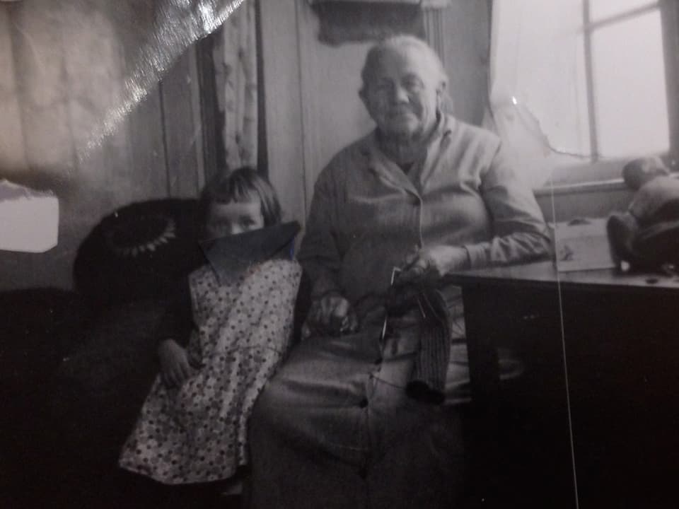 Ein kleines Mädchen steht neben einer alten Frau, die auf einem Stuhl am Tisch sitzt.