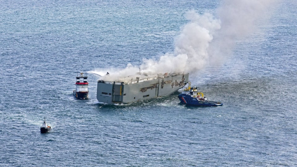 Rauch steigt über dem Frachter im Meer auf, während Löschschiffe auf die Oberseite des Frachters Wasser sprühen.