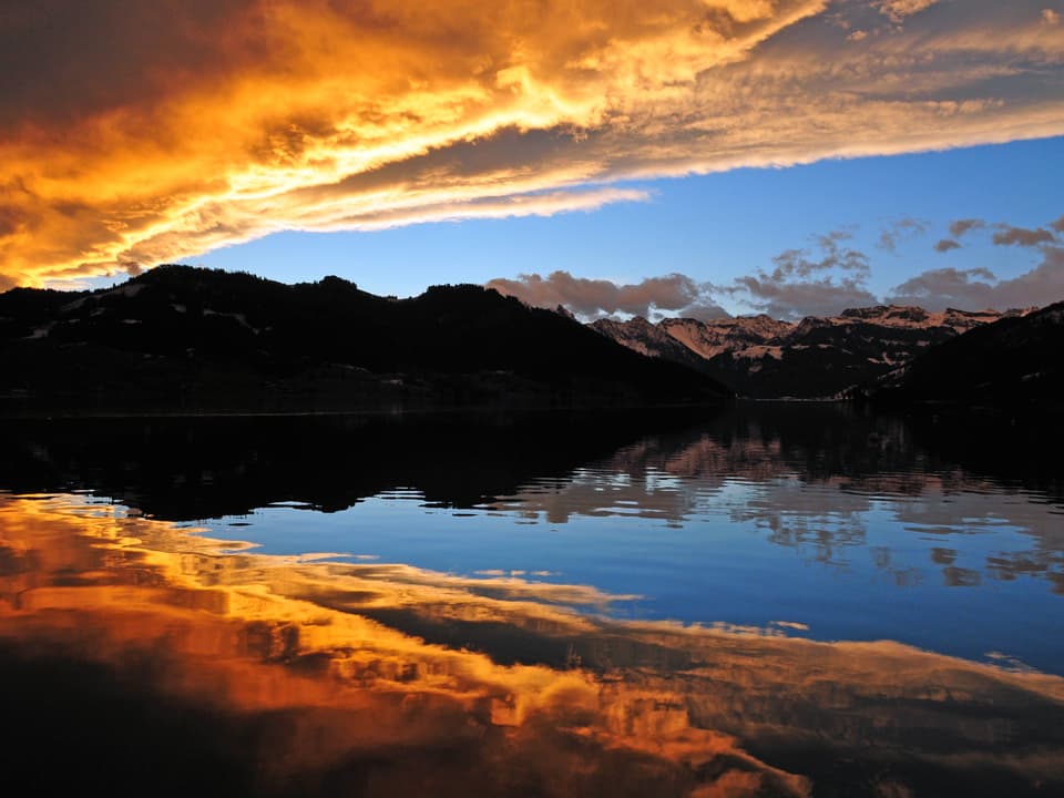 In einem See spiegeln sich die goldenen Wolken und das blaue Föhnfenster. Zwischen dem See und dem Himmel liegt eine dunkle Bergkette.