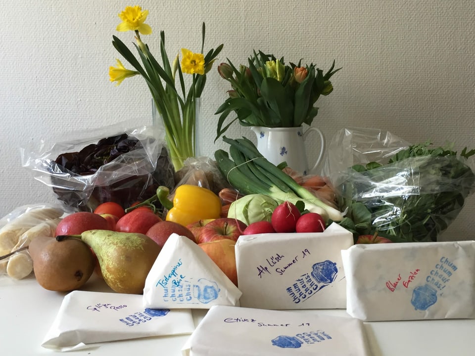 Käse, frisches Obst und Gemüse und im Hintergrund ein Strauss Narzissen und Tulpen liegen auf einem Küchentisch.