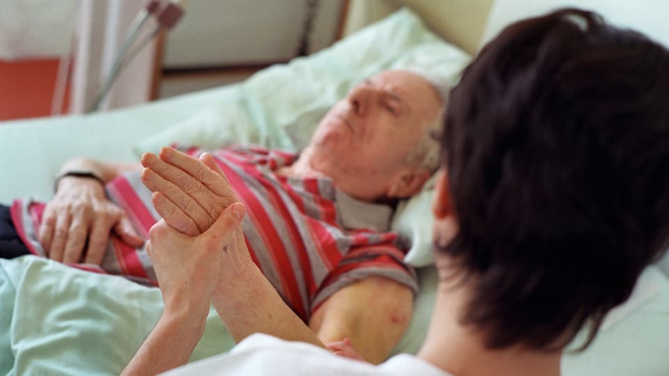 Eine Frau hält die Hand eines älteren Mannes, der in einem Spitalbett liegt. 