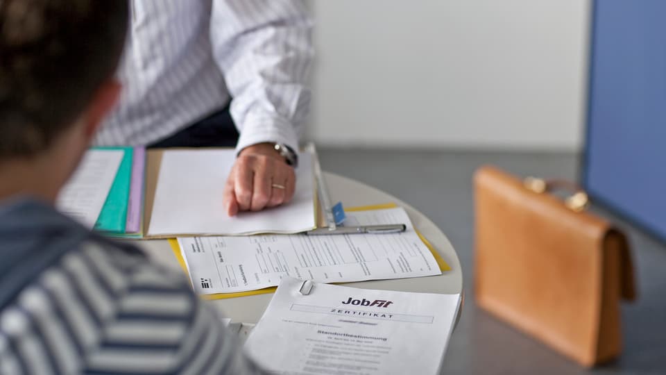 Ein Mann lässt sich auf dem Arbeitslosenamt beraten, vor ihm liegt ein Blatt auf dem Job-Fit steht. Im Hintergrund sieht man unscharf eine braune Mappe.