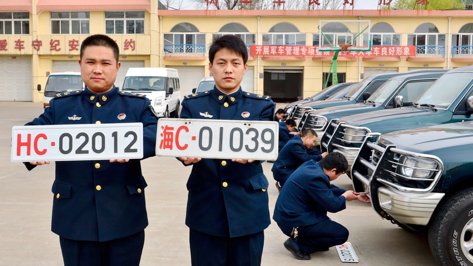 Zwei chinesische Soldaten halen weisse Nummernschilder in den Händen. Im Hintergrund montieren Soldaten Nummernschilder an Autos.