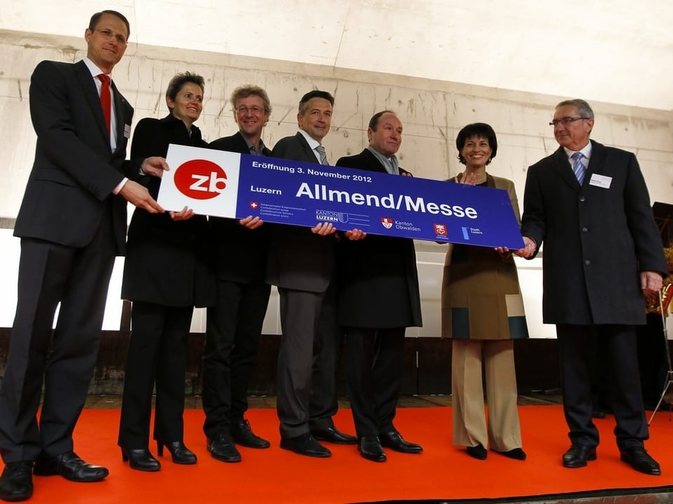Sieben Personen stehen auf einem roten Teppich und halten ein Schild mit der Aufschrift "Luzern Allmend/Messe".
