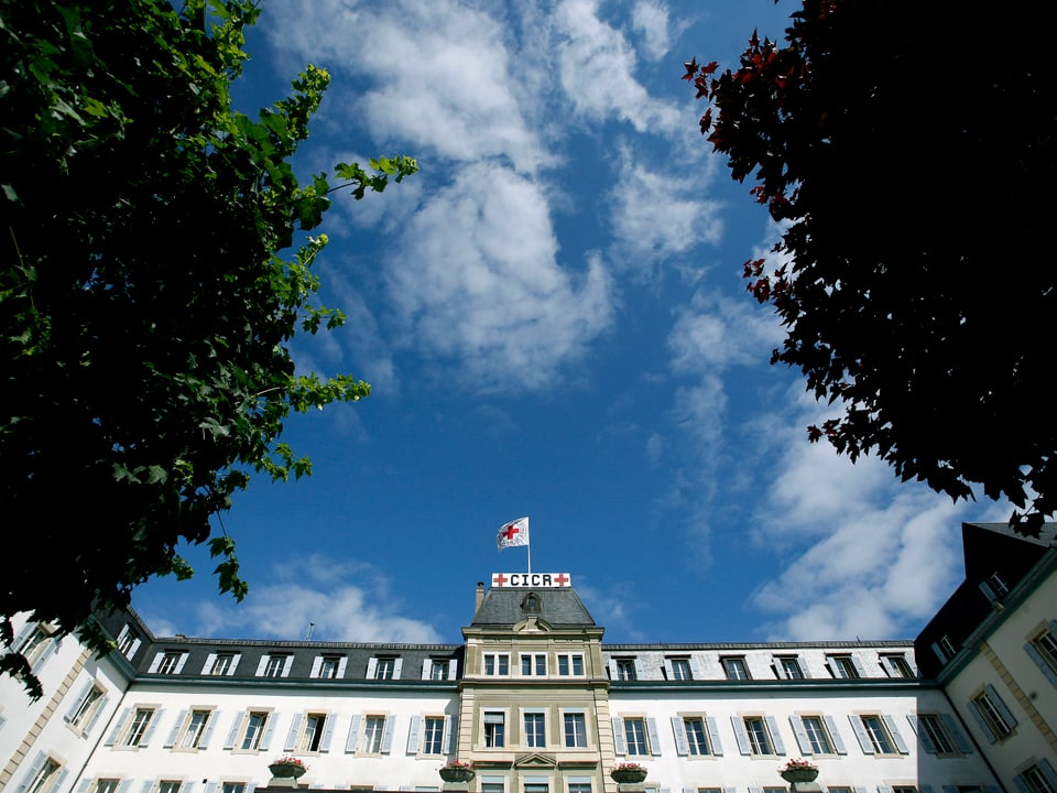 Hauptsitz IKRK, auf dem Dach weht die weisse Flagge mit dem roten Kreuz. Blauer Himmel. Bäume.