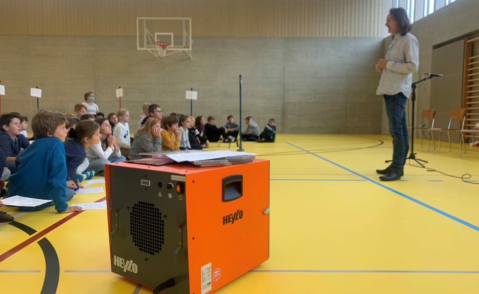 Orange Kiste, Luftreiniger, in einer Turnhalle. Im Hintergrund spricht ein Musiker zu einer Gruppe Kindern.