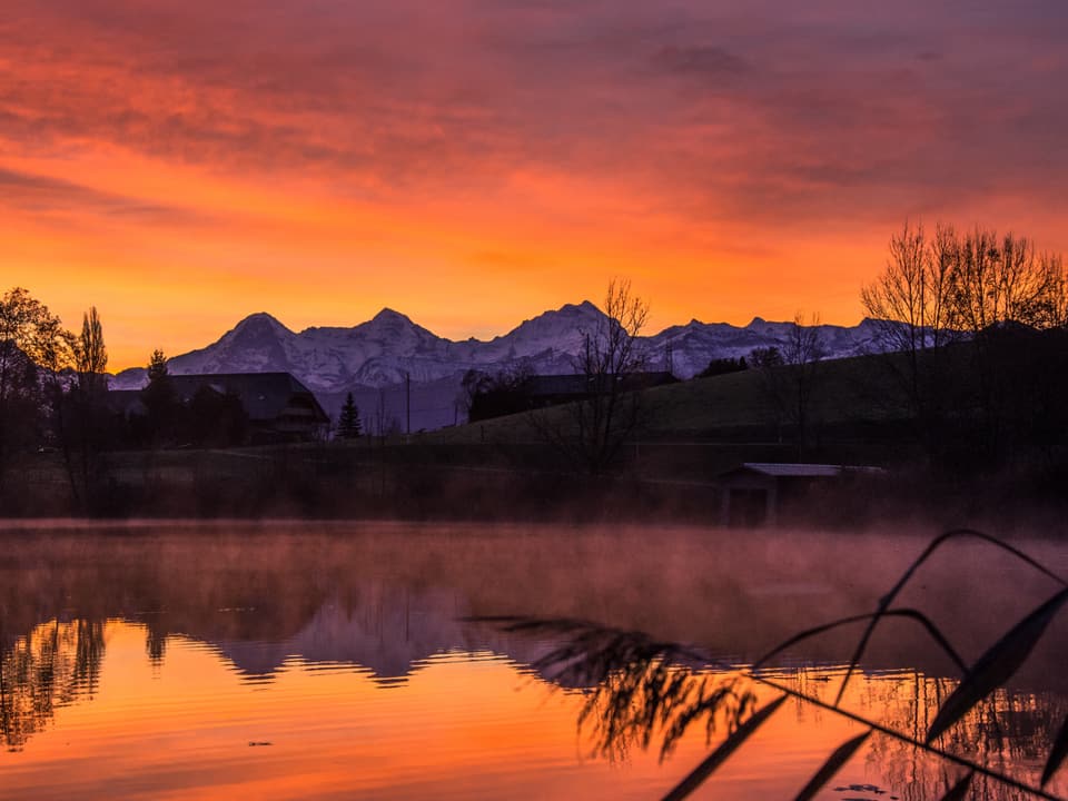 Morgenstimmung an einem See mit orangem Himmel und Schneebergen, die sich im Wasser spiegeln. 
