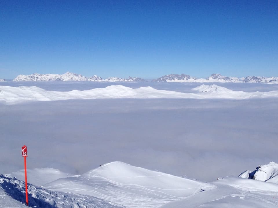 Im Vordergrund eine Skipiste mit Blick über die verschneiten Berggipfel. Die Täler randvoll gefüllt mit Nebel. Darüber stahlblauer Himmel.