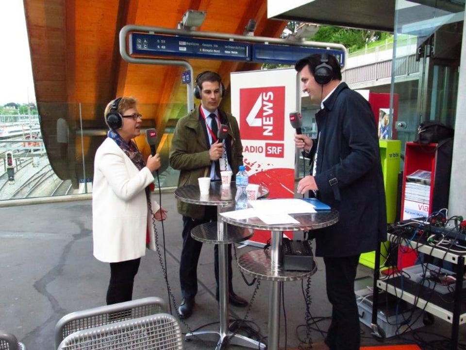 Edith Graf-Litscher und Thierry Burkart stehen an einem Stehtischchen mitten auf der Welle am Bahnhof Bern und sprechen mit Lukas Mäder. Alle tragen Kopfhörer und halten ein Mikrofon in der Hand.