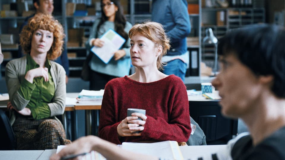 Eine junge Frau sitzt an einem Tisch, um sie herum stehen und sitzen andere Personen. Eine Frau schaut kritisch.