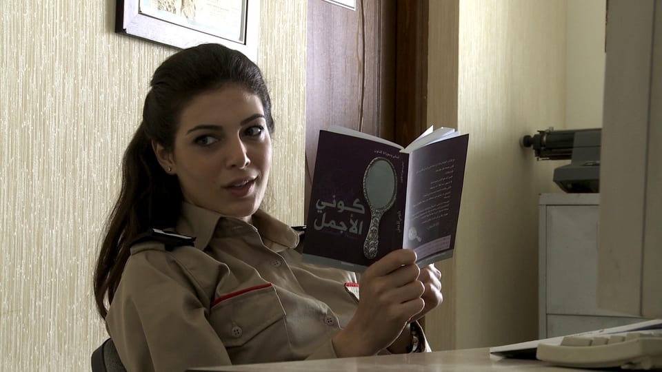 Eine hübsche junge Frau in grüner Uniform sitzt an einem Tisch und schaut von einem Buch auf. Auf dem Cover des Buches ist ein Schminkspiegel zu erkennen.
