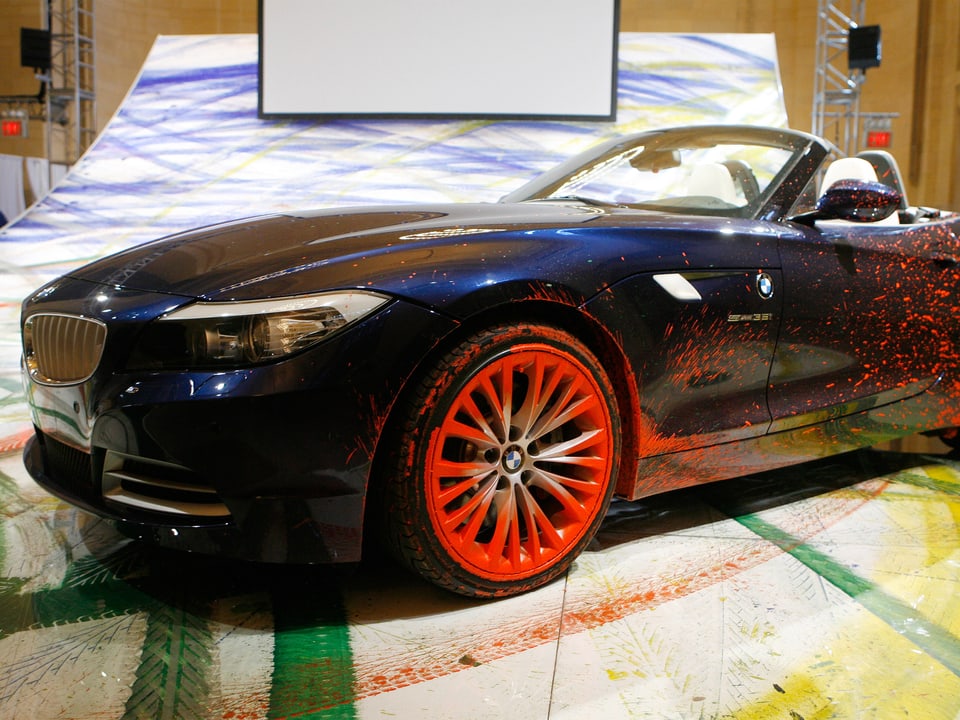 Ein dunkler, farbgekleckerter Sportwagen steht auf einer riesigen Leinwand, auf der mit bunten Reifenspuren übersät ist.