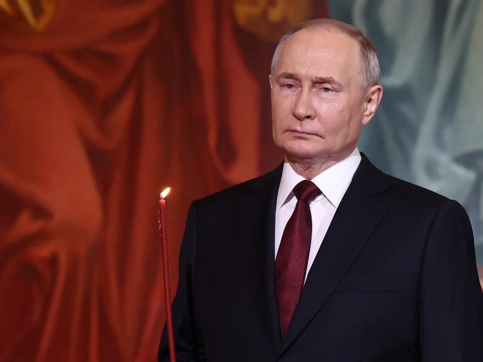 Putin in Anzug hält Kerze vor rotem Vorhang.