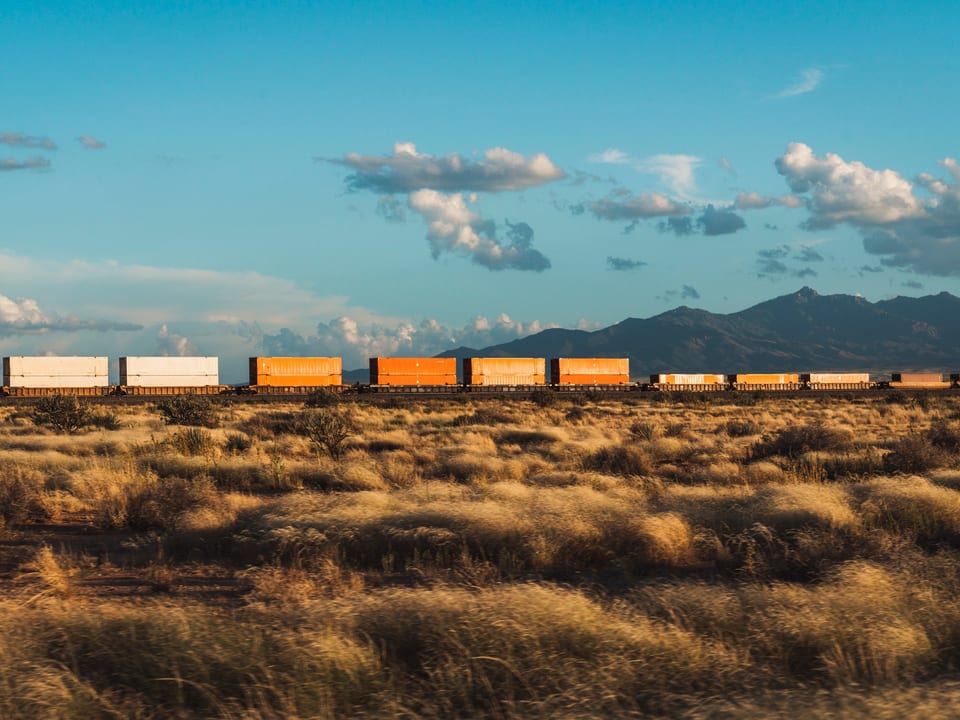 Ein Güterzug in Arizona. Davor eine Wiese. Im Hintergrund sieht man einen Bergzug. 