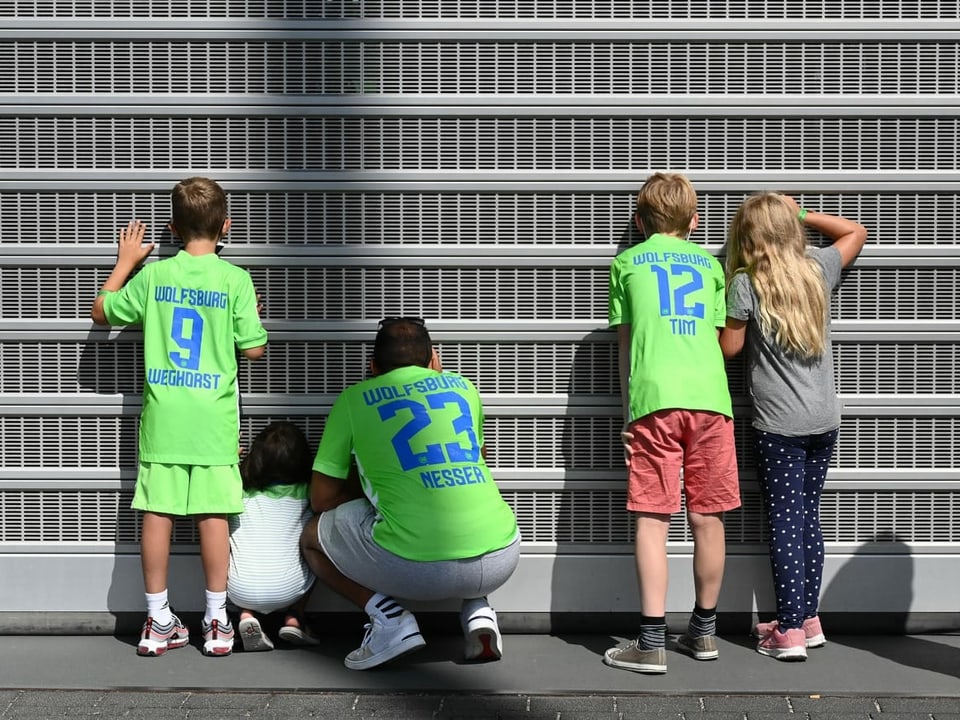 Fünf Personen in Rückenansicht mit Wolfsburg-Trikots blicken durch ein Gitter.