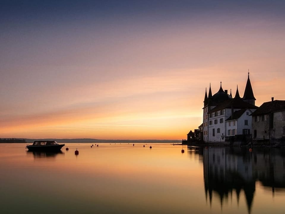 Blick vom Ufer auf glatte Seefläche, in der sich der pastellfarbene Morgenhimmel spiegelt. 