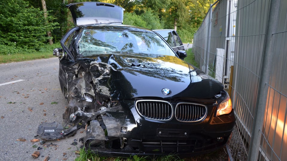 Ein zerstörter BMW-Kombi steht neben der Strasse, an ein Absperrgitter gedrückt.