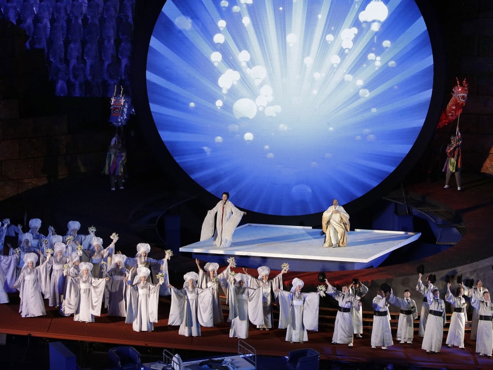 Bühne mit weissgekleideten Menschen und hellem Kreis im Hintergrund.
