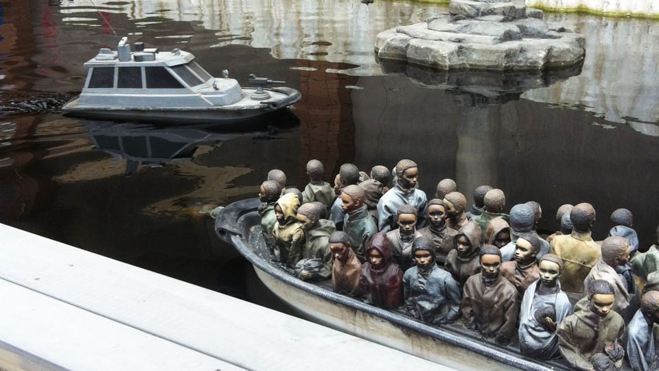 In einem Wasserbecken sind ein Miniatur-Polizeiboot sowie ein Boot voller schwarzer Männer zu sehen. 