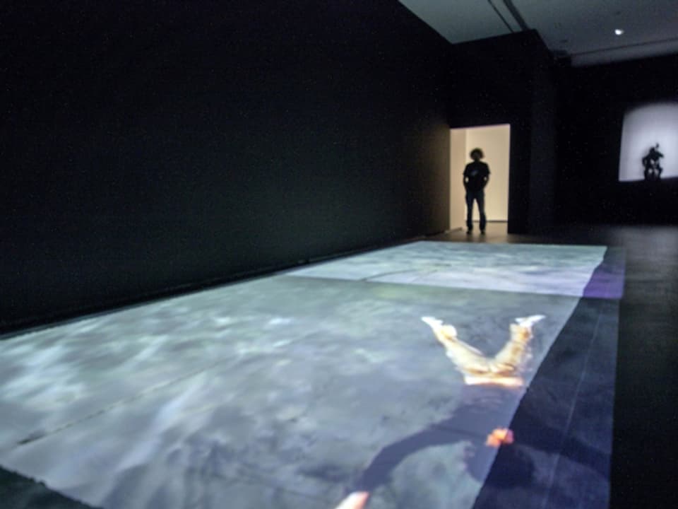 Eine Videoprojektion auf dem Boden, die einen im Wasser treibenden Mann zeigt, davor steht ein Mann.