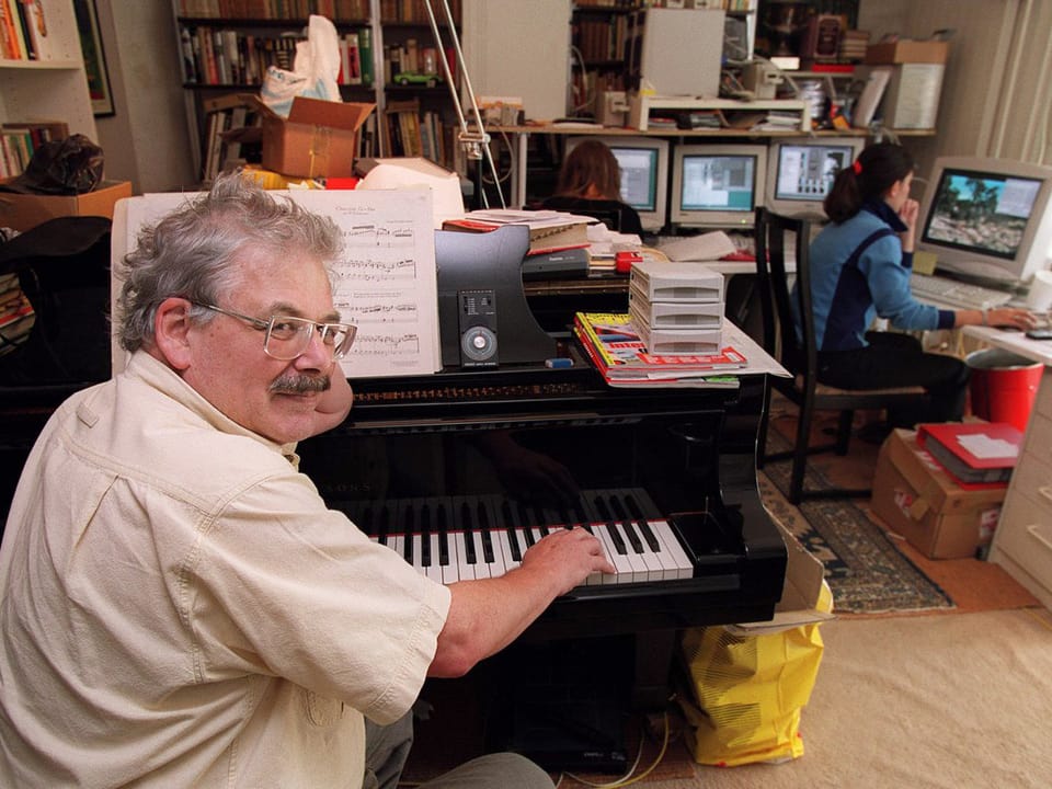 Ein Mann mit Hemd am Klavier, im Hintergrund Frauen an Computern sitzend.