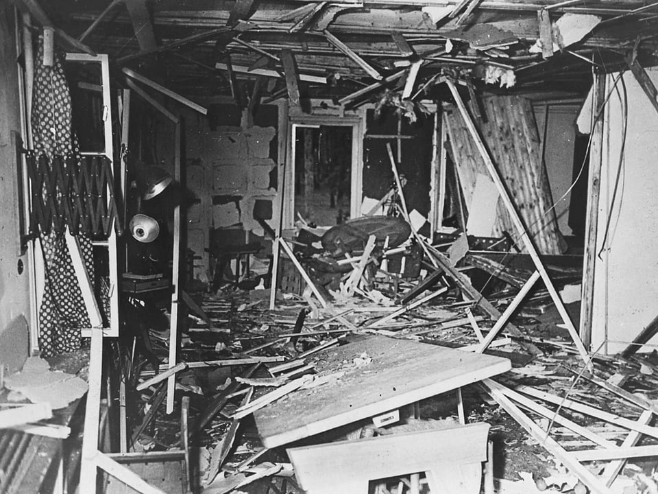 Historisches Schwarzweiss-Bild: Von einer Bombe zerstörter Raum mit kaputtem Tisch und zahlrechen herumliegenden Holzstücken