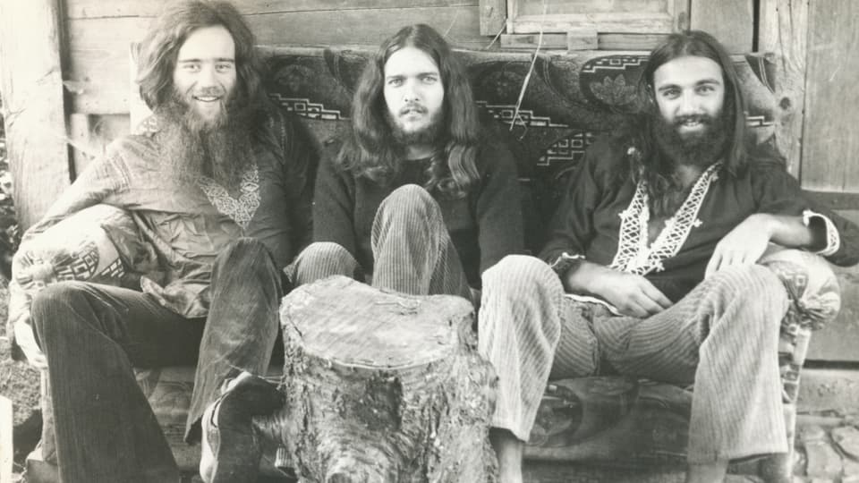 Drei Männer mit langen Haaren und Vollbart sitzen auf einer Bank. 