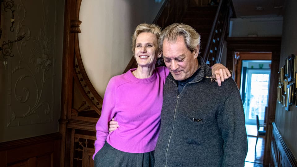 Eine Frau mit kurzen blonden Haaren und violettem Pullover legt ihren Arm um einen Mann mit grauer Jacke.