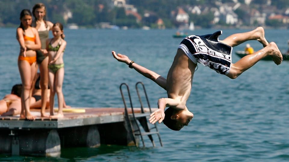 Ein Junge springt in den See, per Salto, auf einem Schwimmfloss schauen Mädchen ihm zu.