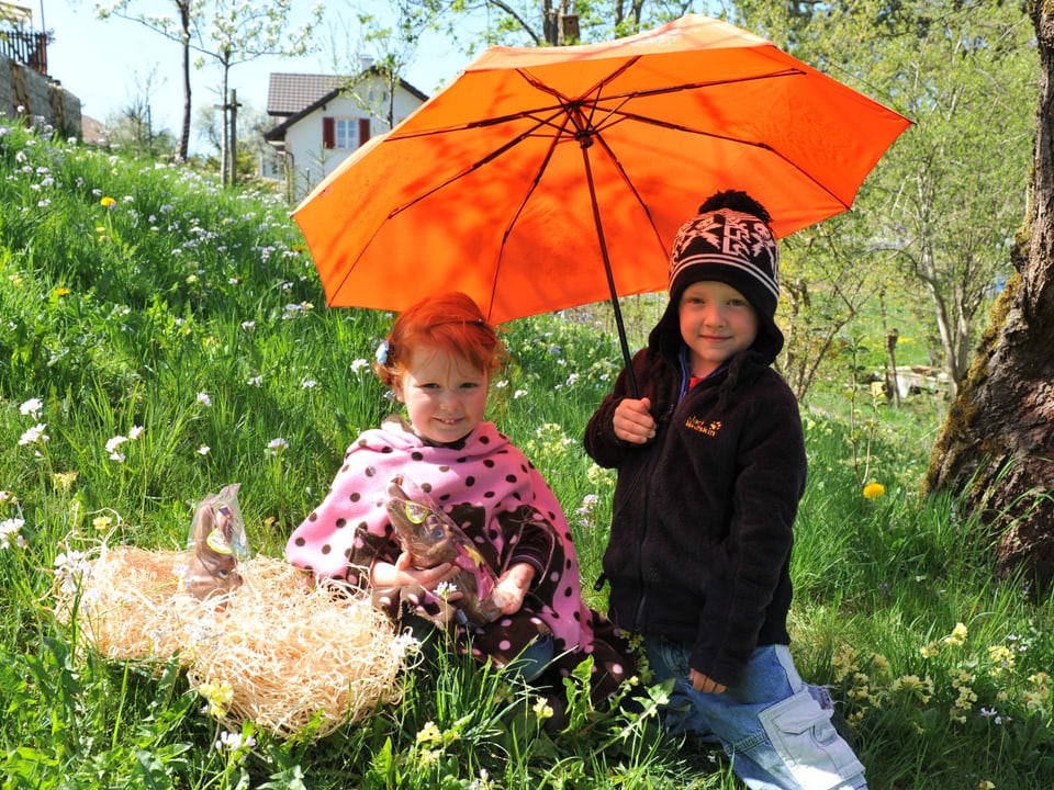 Wiese mit zwei Kindern neben einem Osternest. Der Junge rechts hält einen orangen Sonnenschirm über das Mädchen links. 