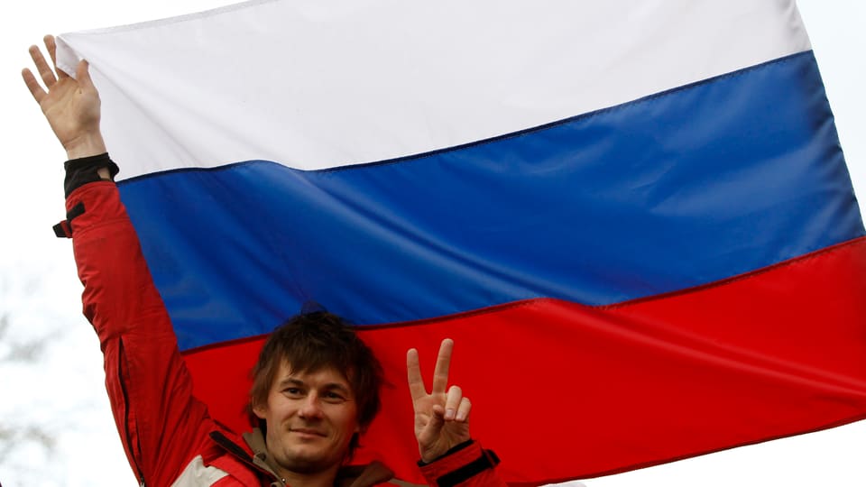 Ein Mann macht mit zwei Fingern ein V-Zeichen vor einer grossen Russland-Fahne.