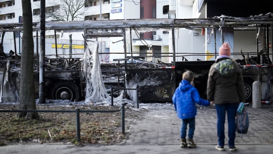 Frau und Kind stehen vor einem ausgebrannten Bus.