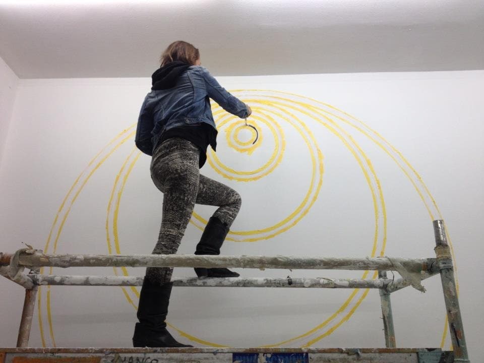 Claudia Comte auf einem Gerüst stehend, auf eine weisse Wand gelbe Kreise malend.