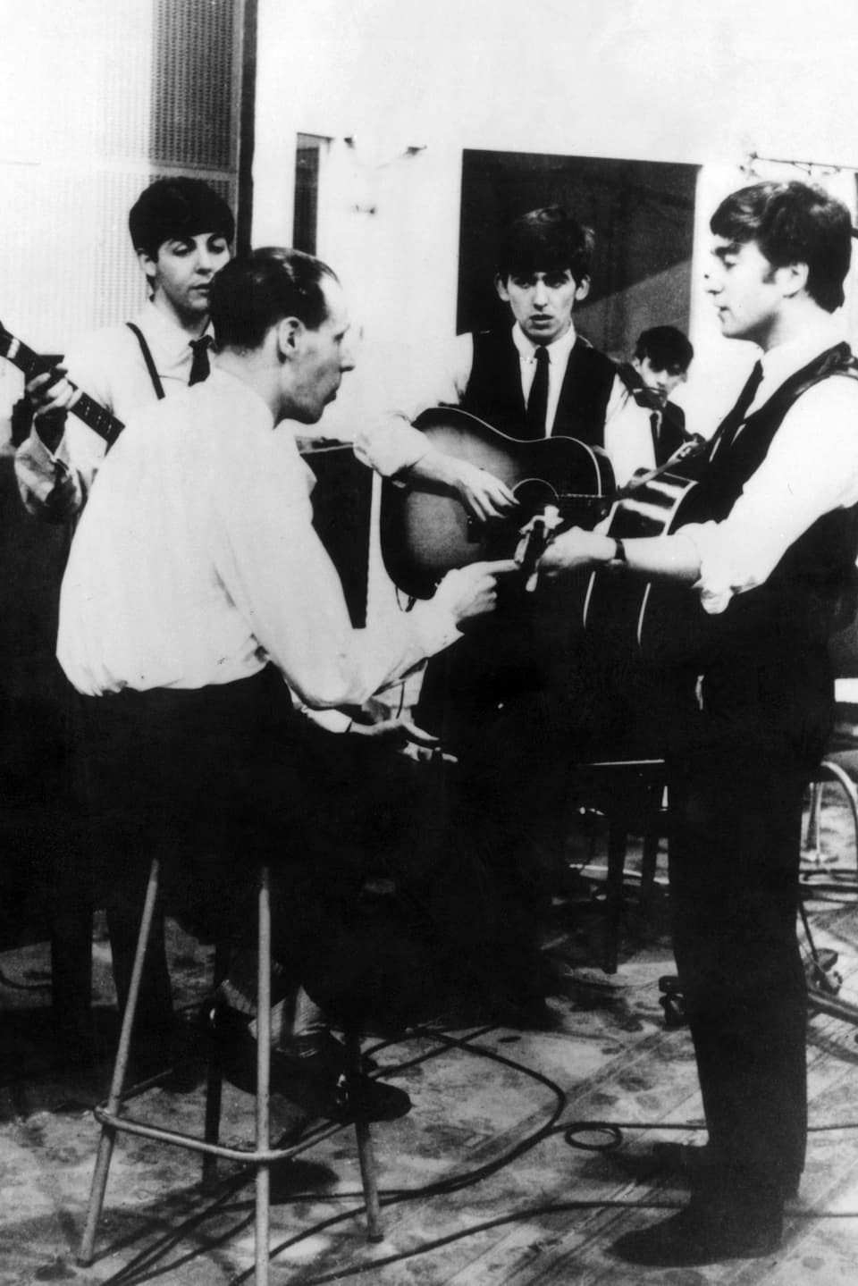 Vier junge Musiker mit ihren Instrumenten, auf einem Hocker sitzt George Martin und gibt Anweisungen.