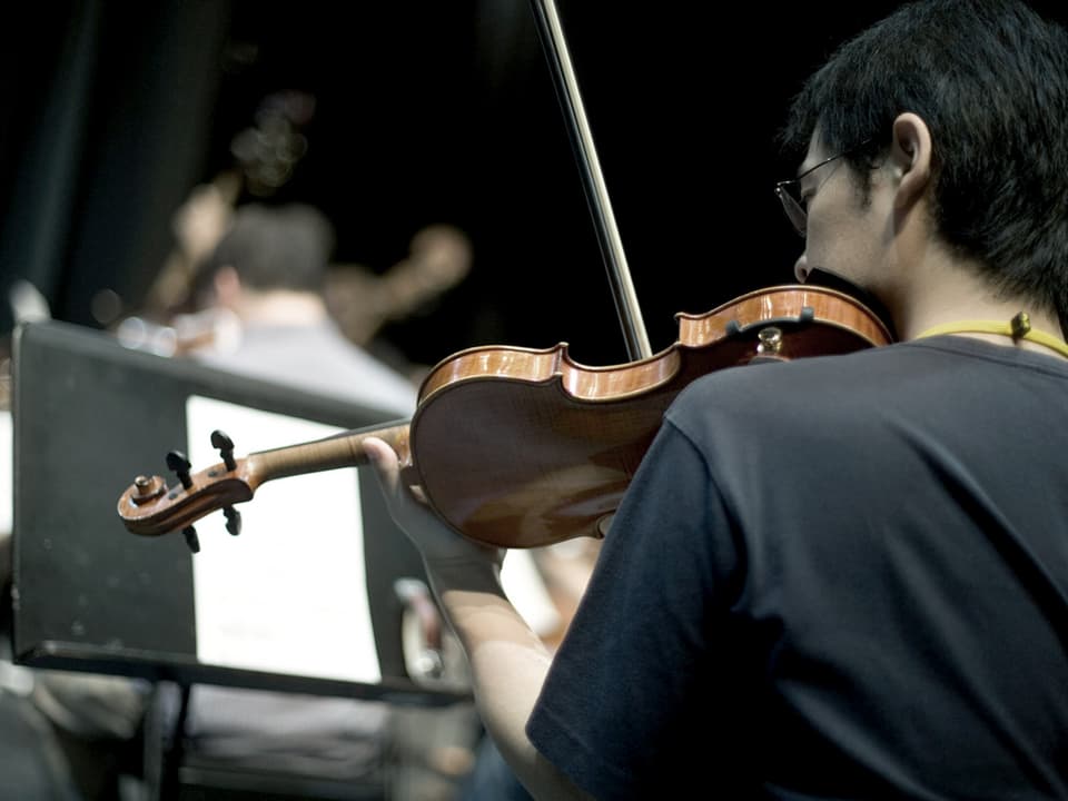 Rückensicht auf einen jungen Mann, der vor einem Notenständer sitzt und Geige spielt. 