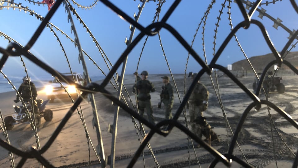 US-Soldaten überprüfen einen Migranten, fotografiert durch einen Maschendrahtzaun.