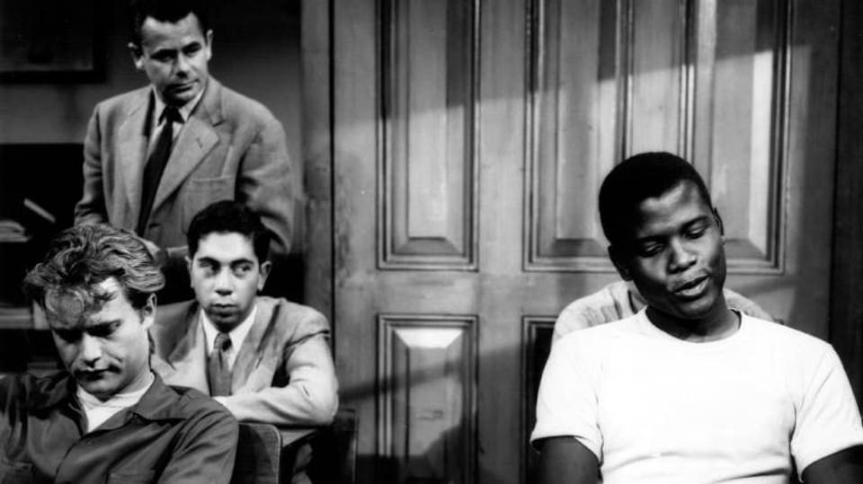 Das schwarzweiss Bild zeigt eine Szene im Klassenzimmer aus dem Film. Links hinten steht Glenn Ford, der den Lehrer Richard Dadier spielt. Die Schüler sitzen auf ihren Plätzen. Drei von ihnen sind auf dem Bild zu sehen.