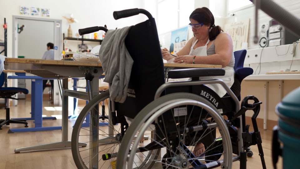 Frau an einem Tisch, daneben ein Rollstuhl