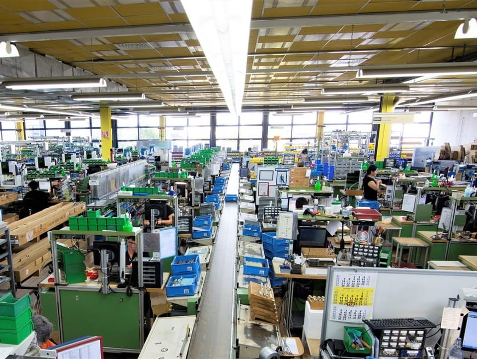 Fabrikhalle mit vielen Arbeitsplätzen und Kisten zur Sortierung von Gegenständen und vereinzelt Personen bei der Arbeit