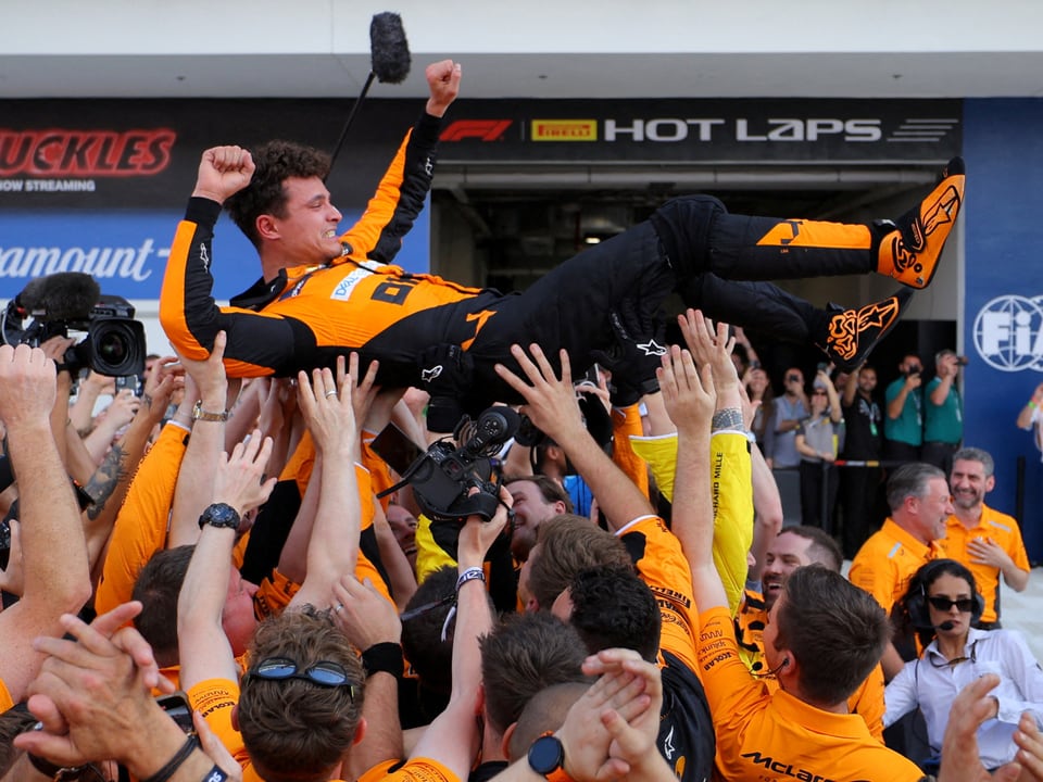 Rennfahrer wird nach einem Sieg von einer Gruppe in orangefarbenen Overalls in der Luft geworfen.