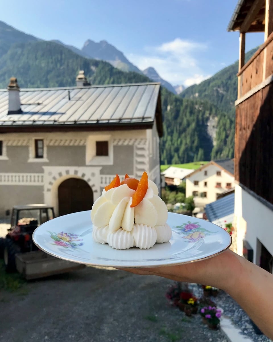 ein gelber Kuchen mit weiss bräunlichen Häubchen darauf. Dahinter eine verschwommene Steingasse, Häuser, blauer Himmel.