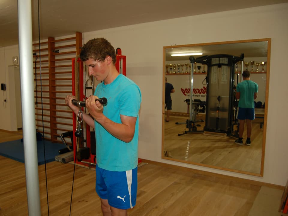 Ein junge Mann mit Hanteln in einem Raum mit verschiedenen Trainingsgeräten.