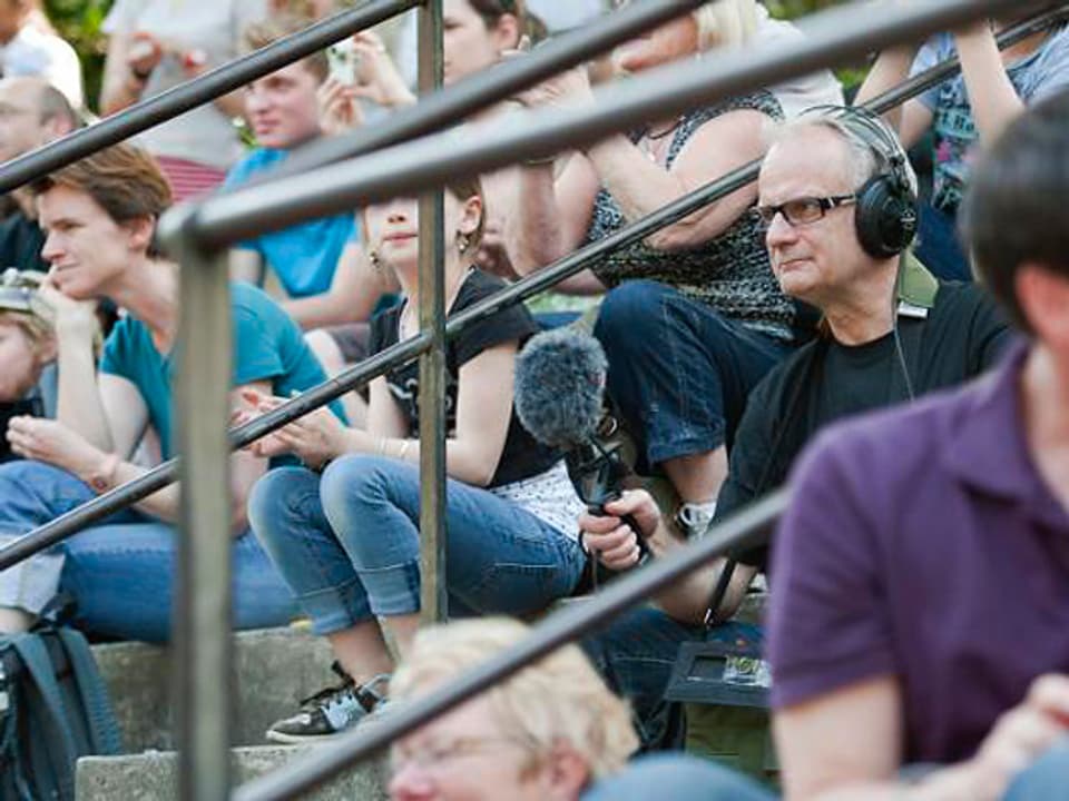 Jack Jakob sitz mit einem Mikrofon und Kopfhörern zwischen anderen Zuschauern auf den Zuschauerrängen bei der Seelöwenfütterung.