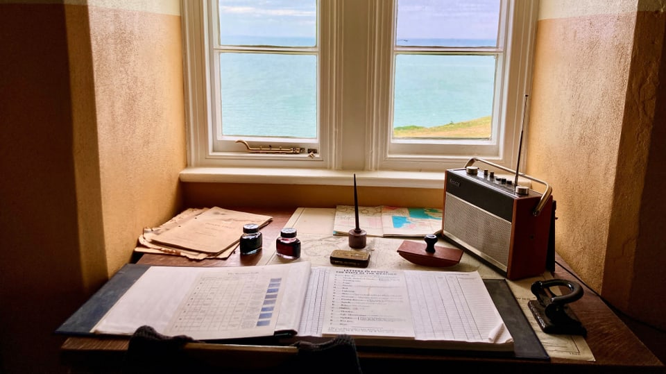 Schreibunterlagen und ein Radio auf einem Schreibtisch in einer Nische am Fenster, Blick auf das Meer. 