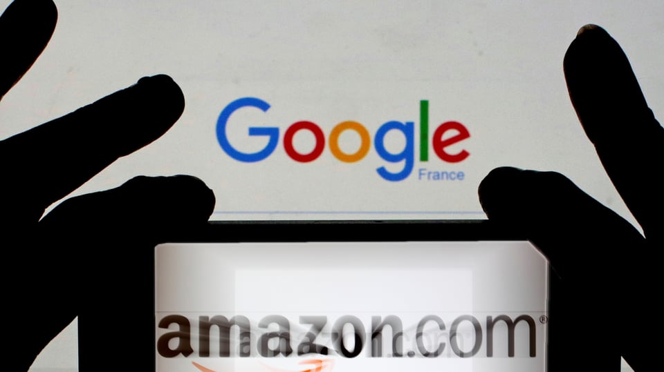 Hände halten ein Smartphone mit dem Amazon-Logo vor einen Bildschirm mit der Google-Suchseite.