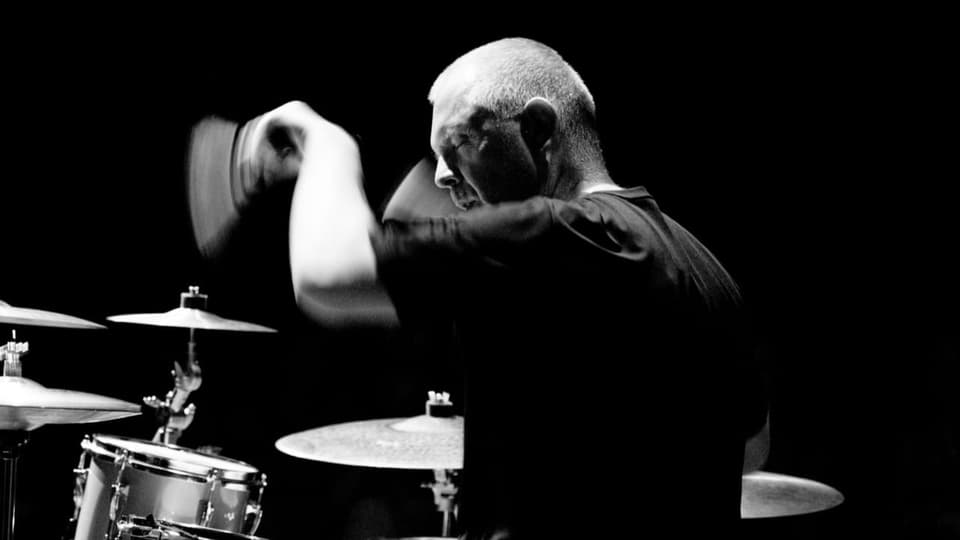 Ein älterer Herr in schwarzem T-Shirt sitzt an einem Schlagzeug; er holt zum Schlag aus, in der Hand hält er Sticks.