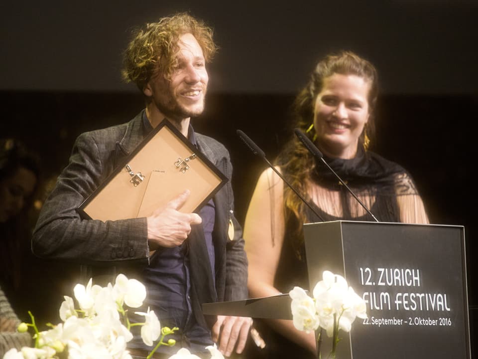 Jan Gassmann am Rednerpult, den Preis in der Hand. Neben ihm eine lächelnde junge Frau.
