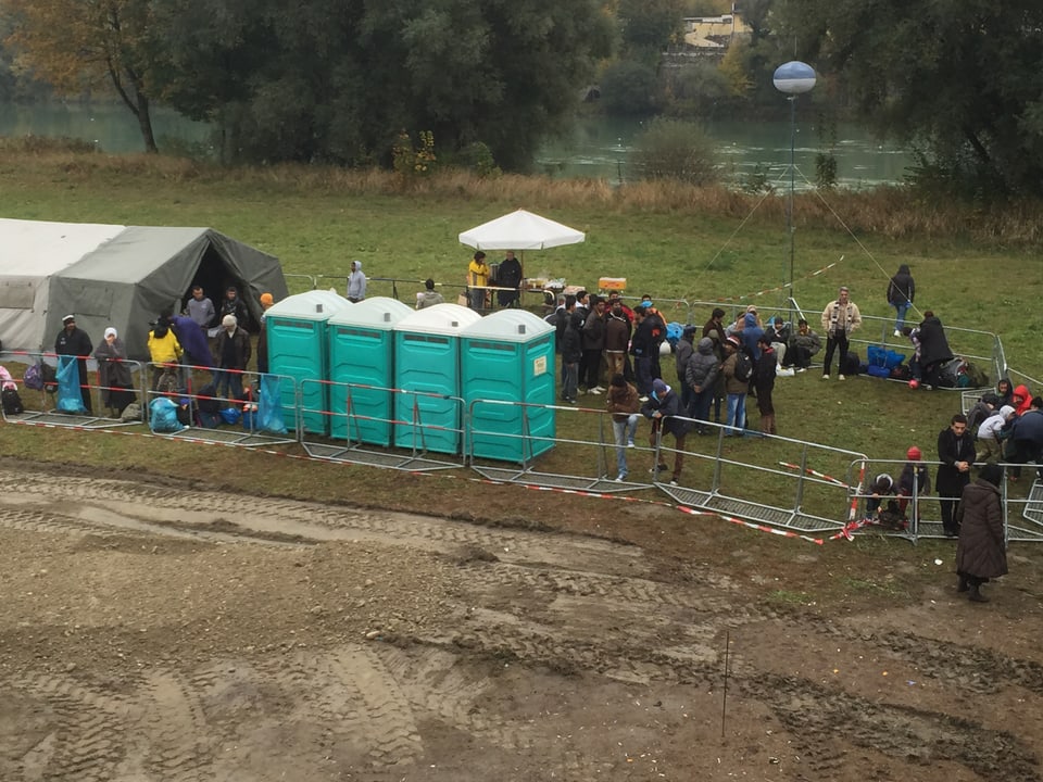 In einem Feld stehen vier mobile Toiletten, ein paar Zelte und Menschen. 