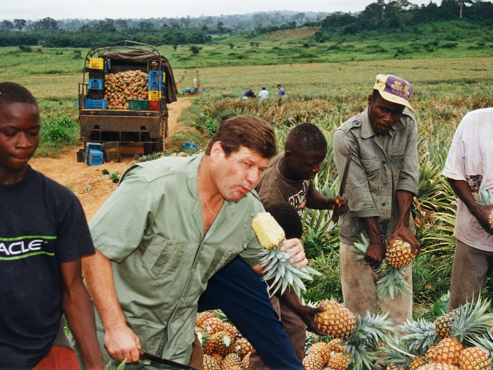 Johann Dähler beisst neben Arbeitern in eine Ananas